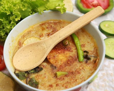 A creamy bowl of soup garnished with herbs, sliced lime, and a green chili, accompanied by a biodegradable spoon. Fresh lettuce, tomato slices, and cucumber slices are arranged around the bowl.