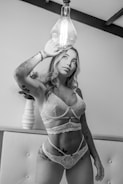 A person wearing lace lingerie stands indoors, touching a large vintage-style light bulb. The setting includes a tufted headboard and a decorative vase in the background. Tattoos adorn the person's arms and legs.