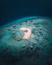 A heart-shaped island surrounded by a network of water villas connected by walkways. The surrounding water ranges from deep blue to turquoise, highlighting the contrast between shallow and deep areas. The island features a sandy area with a small building at its center.