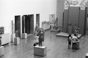 The image depicts a museum interior showcasing various sculptures and artifacts on pedestals. Several visitors are observing the art pieces, which include statues and heads, some of which appear to be of ancient origins. The setting has a clean, modern design with a focus on the exhibits.