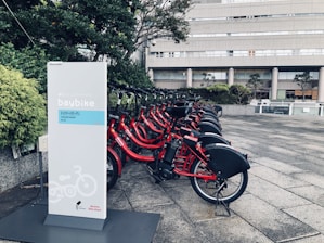 A row of red rental bicycles is parked at a designated bike-sharing station. The bikes are equipped with baskets and are lined up neatly, ready for use. A sign with the words 'baybike' and other text in a foreign language stands prominently in the foreground. The background features an urban setting with a modern building and greenery.