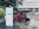 A row of red rental bicycles is parked at a designated bike-sharing station. The bikes are equipped with baskets and are lined up neatly, ready for use. A sign with the words 'baybike' and other text in a foreign language stands prominently in the foreground. The background features an urban setting with a modern building and greenery.