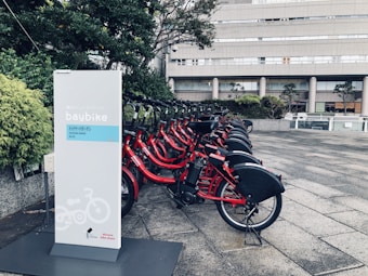 A row of red rental bicycles is parked at a designated bike-sharing station. The bikes are equipped with baskets and are lined up neatly, ready for use. A sign with the words 'baybike' and other text in a foreign language stands prominently in the foreground. The background features an urban setting with a modern building and greenery.