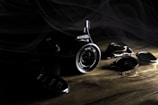 A digital camera with a prominent lens rests on a wooden surface in a dimly lit environment. The brand name is visible on the body of the camera, and several detached lens caps are scattered nearby. Light and shadow interplay creates a dramatic effect, highlighting the camera and its components while smoke or mist partially shrouds the scene.