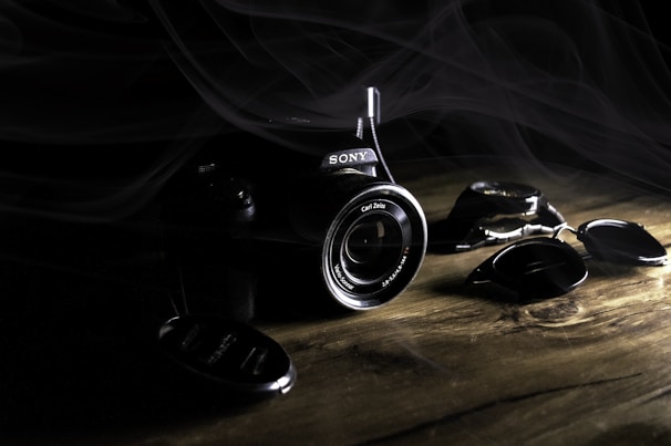 A digital camera with a prominent lens rests on a wooden surface in a dimly lit environment. The brand name is visible on the body of the camera, and several detached lens caps are scattered nearby. Light and shadow interplay creates a dramatic effect, highlighting the camera and its components while smoke or mist partially shrouds the scene.