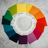 A circular arrangement of Pantone color swatches on a textured gray surface. The swatches form a color wheel with shades transitioning from greens to blues, purples, reds, oranges, and yellows. Each swatch is labeled with its respective Pantone number.