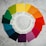 A circular arrangement of Pantone color swatches on a textured gray surface. The swatches form a color wheel with shades transitioning from greens to blues, purples, reds, oranges, and yellows. Each swatch is labeled with its respective Pantone number.