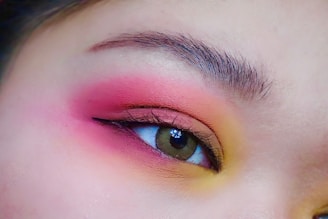 A close-up of a human eye with vibrant makeup, featuring a gradient of pink, red, and yellow eyeshadow around the eyelid. The eyebrow is well-defined with natural hair details.