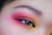 A close-up of a human eye with vibrant makeup, featuring a gradient of pink, red, and yellow eyeshadow around the eyelid. The eyebrow is well-defined with natural hair details.