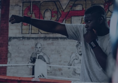 A person practicing a boxing stance in a gym setting, with a mural of a boxer on the brick wall in the background. The individual is wearing a gray shirt and has a focused expression, with their fist extended in a punching motion.