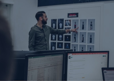 A man in a green shirt points at a board with various printed designs, possibly displaying water bottle mockups. Computer monitors with spreadsheet and email applications appear in the foreground, suggesting an office or design studio setting.