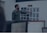A man in a green shirt points at a board with various printed designs, possibly displaying water bottle mockups. Computer monitors with spreadsheet and email applications appear in the foreground, suggesting an office or design studio setting.