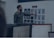 A man in a green shirt points at a board with various printed designs, possibly displaying water bottle mockups. Computer monitors with spreadsheet and email applications appear in the foreground, suggesting an office or design studio setting.