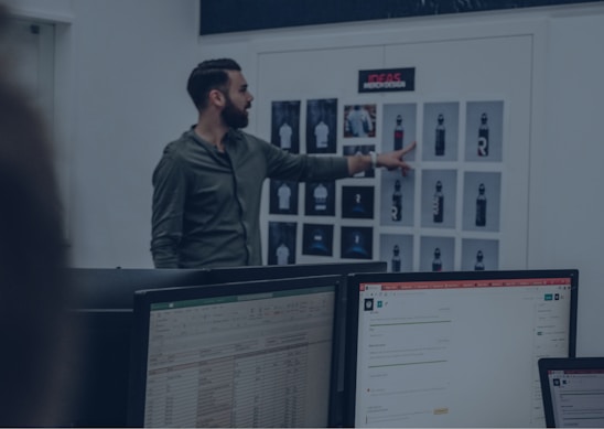 A man in a green shirt points at a board with various printed designs, possibly displaying water bottle mockups. Computer monitors with spreadsheet and email applications appear in the foreground, suggesting an office or design studio setting.