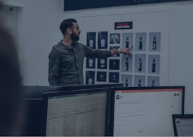 A man in a green shirt points at a board with various printed designs, possibly displaying water bottle mockups. Computer monitors with spreadsheet and email applications appear in the foreground, suggesting an office or design studio setting.