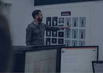 A man in a green shirt points at a board with various printed designs, possibly displaying water bottle mockups. Computer monitors with spreadsheet and email applications appear in the foreground, suggesting an office or design studio setting.