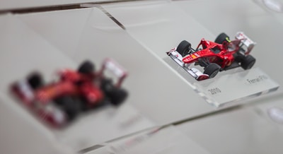 A detailed scale model of a red Formula 1 race car, displayed on a clear angled platform with the label '2010 Ferrari F10'. Another blurred model is visible in the background, highlighting the focus on the sharp and intricate design of the foreground car.