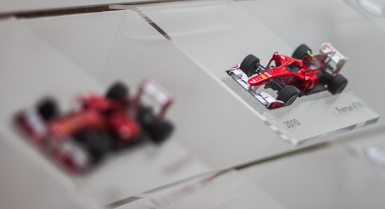 A detailed scale model of a red Formula 1 race car, displayed on a clear angled platform with the label '2010 Ferrari F10'. Another blurred model is visible in the background, highlighting the focus on the sharp and intricate design of the foreground car.