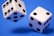 Two white dice with black dots displayed on a vibrant blue background. Both dice are in mid-air, showing the dynamic motion and interaction between the objects.