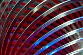 Curved metal wires are arrayed closely together, creating a series of parallel arcs against a background of vibrant red and blue striped patterns. The reflective surface of the metal arcs captures and diffuses the colorful lights, adding a dynamic and abstract aesthetic.