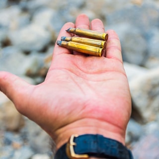 A person's hand is holding three bullets. The background is blurred, featuring a rocky surface.