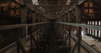 A dark and abandoned industrial structure with metal beams and a symmetrical perspective. The worn-out wooden ceiling and support beams create a sense of neglect and decay. Dust and debris are scattered across the floor, and the lighting is dim, adding to the somber atmosphere.