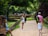 People are walking along a brick pathway surrounded by lush green trees and ancient brick structures. A woman with sunglasses and a backpack is holding a camera, while others are dressed casually, enjoying the outdoor setting.