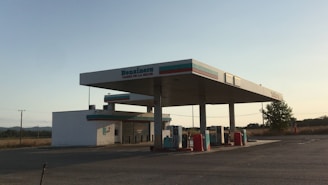 A gas station located in a remote and open area with clear skies. The structure consists of a main fueling area with several gas pumps and a small building that likely serves as a convenience store. There are no vehicles or people present in the image. The surroundings include some trees and distant hills.