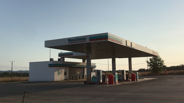 A gas station located in a remote and open area with clear skies. The structure consists of a main fueling area with several gas pumps and a small building that likely serves as a convenience store. There are no vehicles or people present in the image. The surroundings include some trees and distant hills.