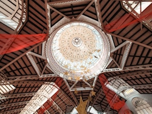 An architectural dome with intricate designs, featuring a circular skylight surrounded by decorative patterns and geometric shapes. The structure includes metal beams and glass panes. Red fabric drapes hang from the ceiling, adding a splash of color and dynamic movement.