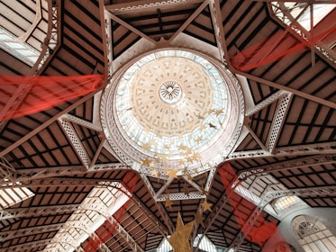 An architectural dome with intricate designs, featuring a circular skylight surrounded by decorative patterns and geometric shapes. The structure includes metal beams and glass panes. Red fabric drapes hang from the ceiling, adding a splash of color and dynamic movement.