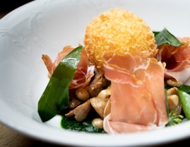 A gourmet dish with a crispy fried ball, slices of cured meat, mushrooms, and a vibrant green sauce. The presentation is refined, with ingredients artistically arranged in a white, textured bowl.