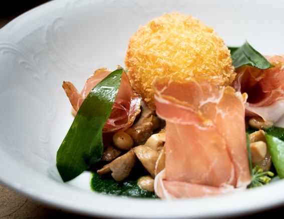 A gourmet dish with a crispy fried ball, slices of cured meat, mushrooms, and a vibrant green sauce. The presentation is refined, with ingredients artistically arranged in a white, textured bowl.