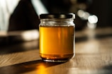 A glass jar filled with golden honey is placed on a wooden table. The lighting creates a warm, inviting atmosphere with soft shadows and highlights that enhance the richness of the honey's color.