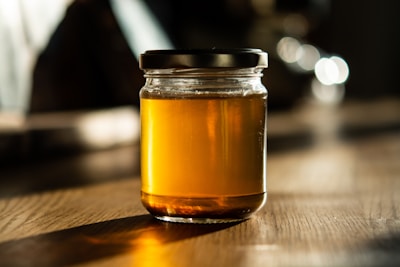 A glass jar filled with golden honey is placed on a wooden table. The lighting creates a warm, inviting atmosphere with soft shadows and highlights that enhance the richness of the honey's color.