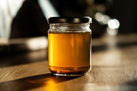 A glass jar filled with golden honey is placed on a wooden table. The lighting creates a warm, inviting atmosphere with soft shadows and highlights that enhance the richness of the honey's color.