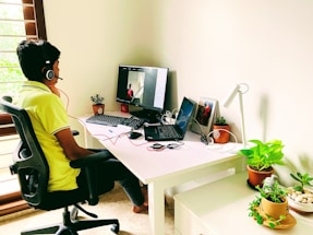 A person with short hair is sitting on an office chair, wearing a headset and a yellow shirt, while facing a computer screen. The desk is organized with two laptops, a keyboard, several potted plants, a desk lamp, and some electronics. The room has natural lighting coming from a window with wooden slats.