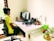 A person with short hair is sitting on an office chair, wearing a headset and a yellow shirt, while facing a computer screen. The desk is organized with two laptops, a keyboard, several potted plants, a desk lamp, and some electronics. The room has natural lighting coming from a window with wooden slats.