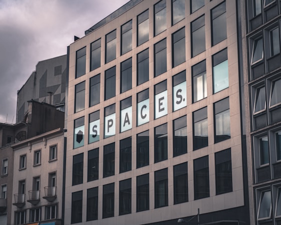 A multi-story building with large rectangular windows. The facade has a modern design with a sign displaying the word 'SPACES'. The sky is overcast, giving the scene a somewhat dim lighting.