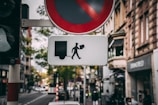 An urban street scene with a focus on a unique directional sign. The sign depicts a stylized figure of a man with a hat and a suitcase walking towards a square shape. Above it is a no-stopping traffic sign. In the background, there are buildings, trees, and several cars and pedestrians, giving a lively city atmosphere.