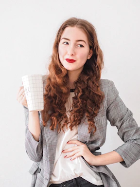 A person with long, curly red hair is holding a large grid-patterned mug. They wear a plaid blazer over a white shirt and have red lipstick and painted nails. The pose is casual and thoughtful, set against a simple white background.