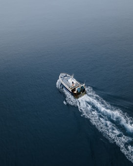Aerial view of a fishing boat cutting through calm, expansive blue ocean waters, leaving a trail of white frothy waves behind.