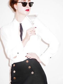 A person dressed in a stylish outfit, wearing a white shirt with black tie detail and a black skirt with circular accents, is holding a martini glass. They have red lipstick, nail polish, and are wearing dark sunglasses.