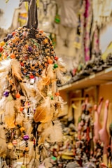 An intricately designed dreamcatcher decorated with colorful beads and feathers. The background features blurred decorative items and souvenirs, suggesting a shop or market setting.