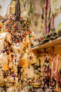 An intricately designed dreamcatcher decorated with colorful beads and feathers. The background features blurred decorative items and souvenirs, suggesting a shop or market setting.