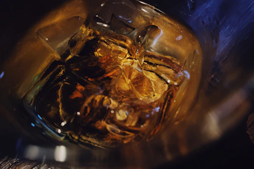 Close-up of a whiskey glass glowing softly on a wooden bar with amber liquid and ice.
