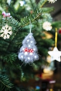 A close-up of a Christmas tree branch adorned with handmade ornaments, including a grey felt figure with white dots and small red mushrooms, and a straw star hanging nearby. The background features a blurred array of green pine needles and soft bokeh lights.