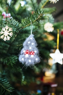 A close-up of a Christmas tree branch adorned with handmade ornaments, including a grey felt figure with white dots and small red mushrooms, and a straw star hanging nearby. The background features a blurred array of green pine needles and soft bokeh lights.