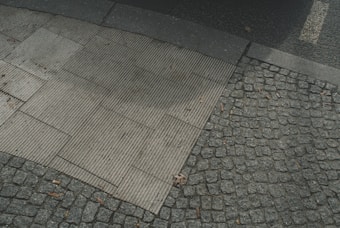 A close-up view of a sidewalk intersection with two distinct paving patterns. One side features rectangular, ribbed concrete slabs, while the other displays a cobblestone texture. The pavement is littered with small twigs and leaves, and there are subtle shadows cast across the stones.