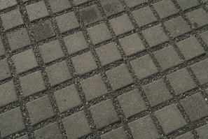 A close-up view of a tiled pavement featuring evenly spaced square concrete tiles separated by small gravel or pebbles, creating a grid pattern.